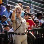 A woman emcee speaks to a crowd in bleachers, pointing and engaging during an outdoor event.