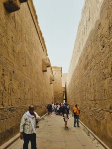 Visitors walk through a corridor of an ancient Egyptian temple with hieroglyphic carvings.