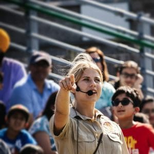 A woman emcee speaks to a crowd in bleachers, pointing and engaging during an outdoor event.